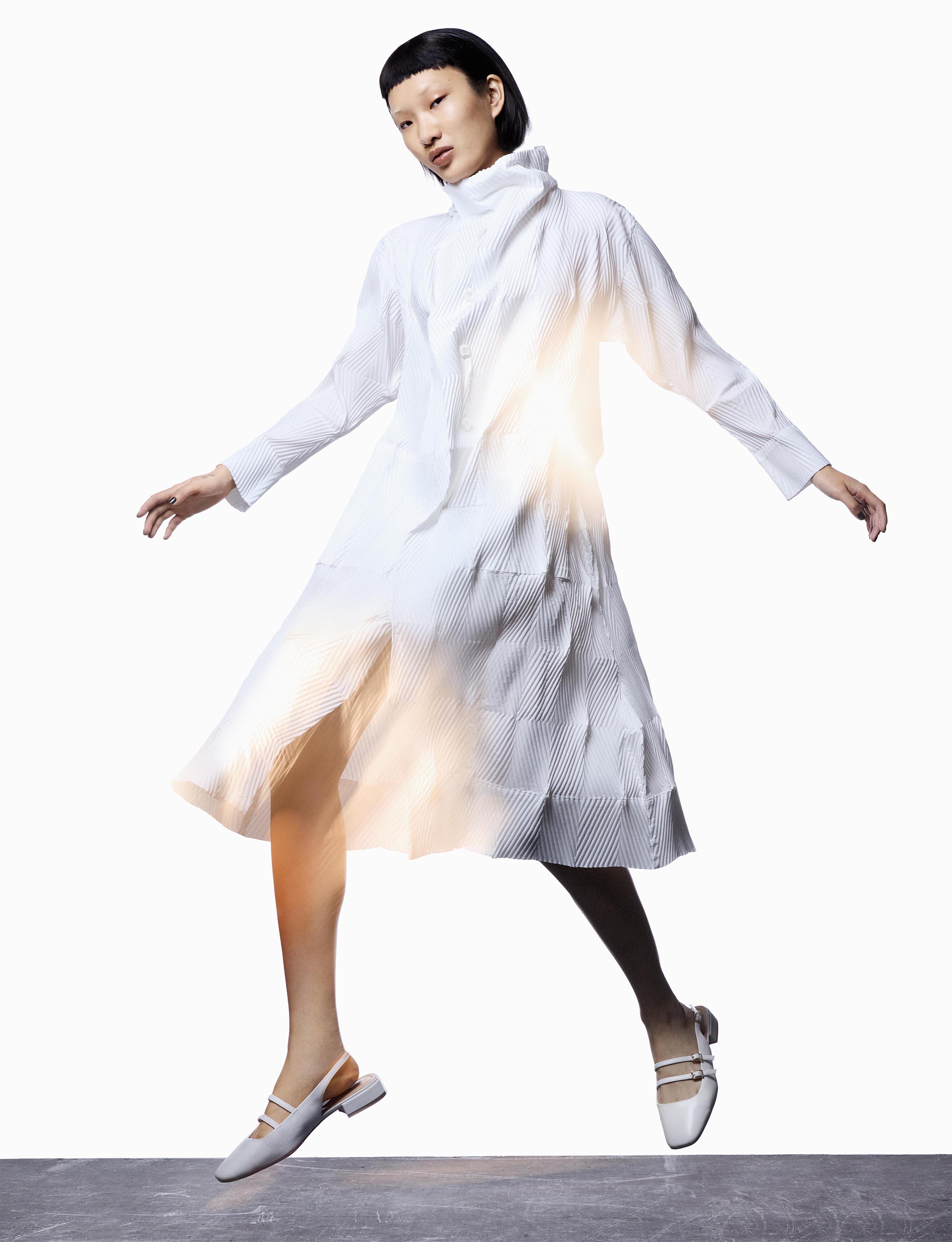 Full-length shot of a stylish Asian female model in a pleated white dress and matching shoes, dynamically posed against a white backdrop.