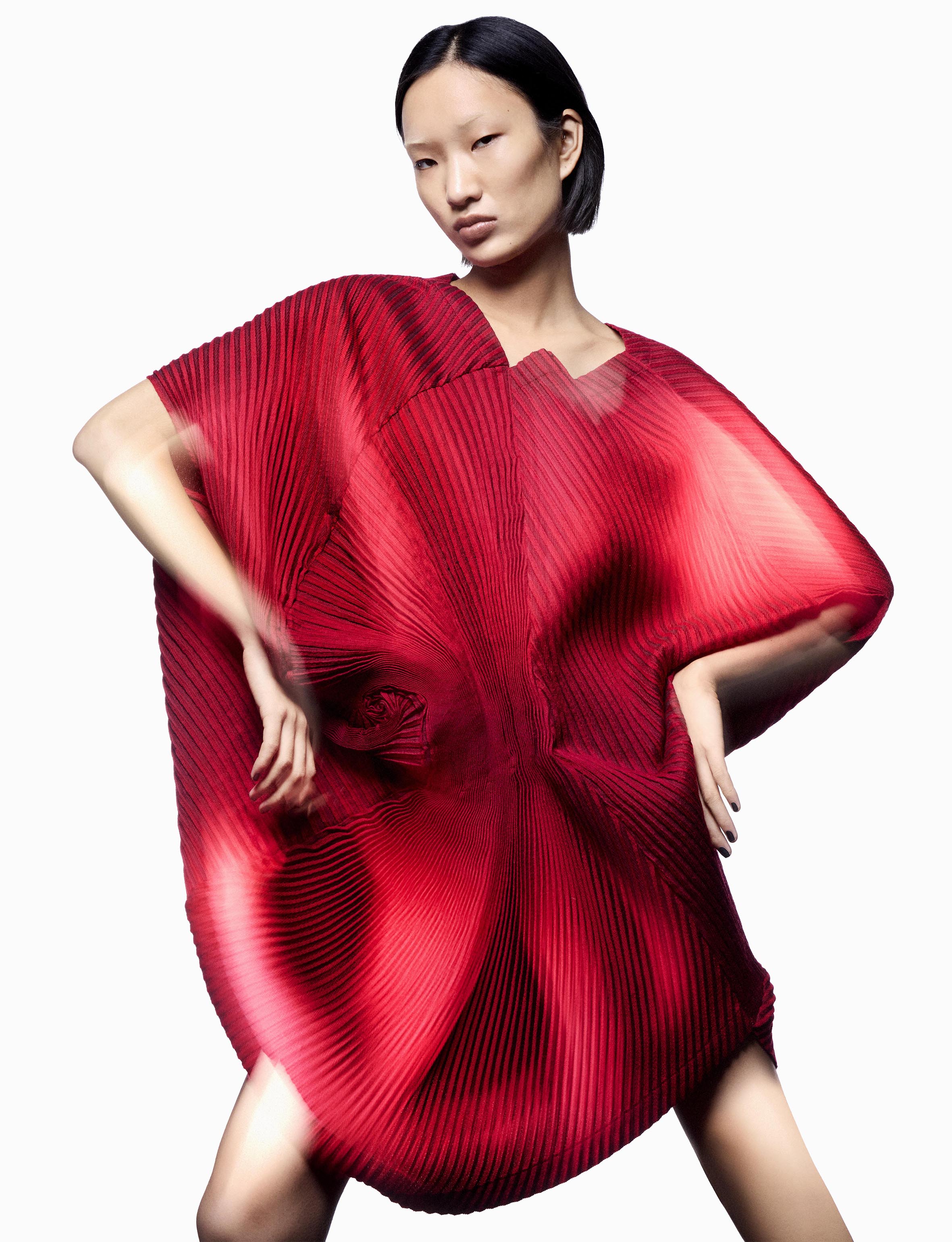 Asian female fashion model wears a flowing, vibrant red dress with a textured, pleated fabric against a bright white studio background.