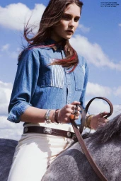 Woman in denim shirt wearing silver bracelets riding a horse against a clear sky with scattered clouds