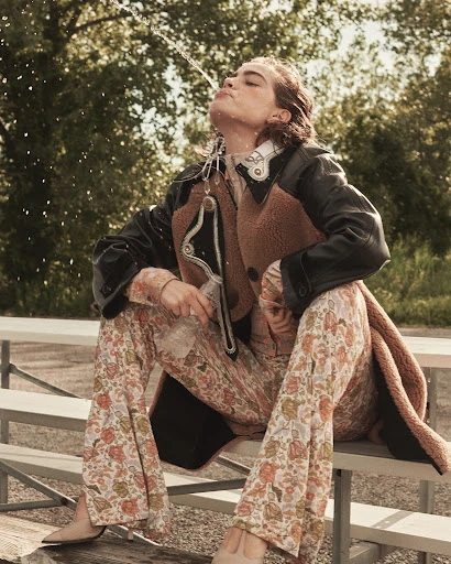 A young woman in fashionable clothing sitting on a metal bench drinking water from a fountain in an outdoor setting with trees.
