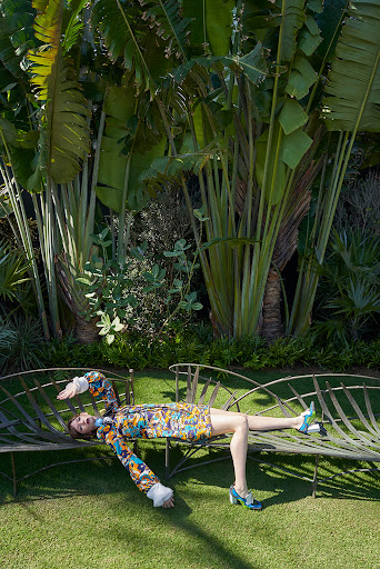 Woman in patterned dress and heels lies on a bench in a lush green garden with tall tropical plants and trees in the background.