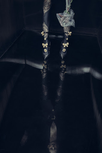 Fashionable woman's legs and feet in decorative high heels are reflected on a shiny black floor in a dark, moody indoor setting.