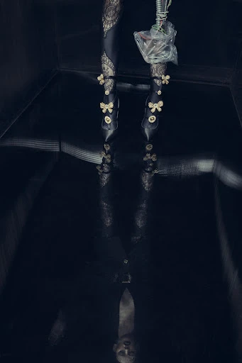 Fashionable woman's legs and feet in decorative high heels are reflected on a shiny black floor in a dark, moody indoor setting.