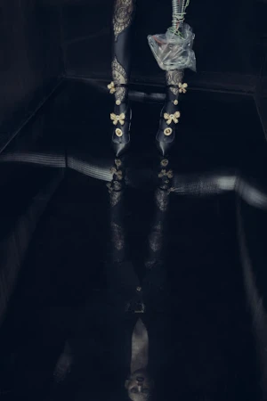 Fashionable woman's legs and feet in decorative high heels are reflected on a shiny black floor in a dark, moody indoor setting.