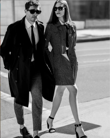 Black and white portrait of a man in a dark trench coat and a woman in a patterned coat walking outdoors