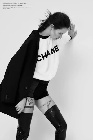 Model in white sweater and dark coat leaning against white wall wearing latex boots