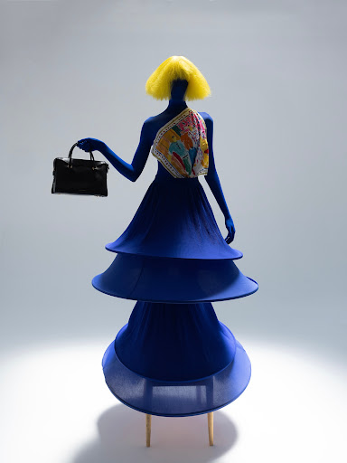 Abstract mannequin wearing a blue sculptural dress and yellow wig holds a black purse against a white backdrop in a studio shot.