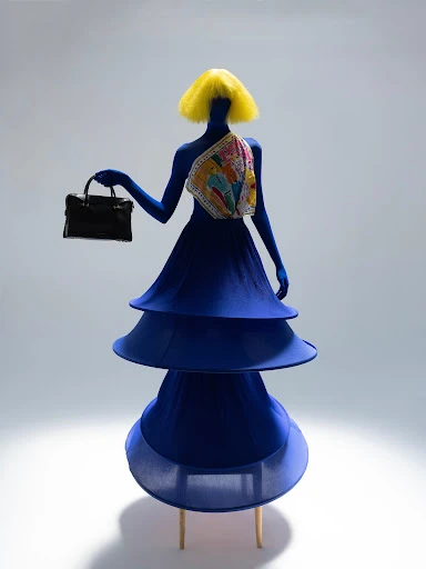 Abstract mannequin wearing a blue sculptural dress and yellow wig holds a black purse against a white backdrop in a studio shot.