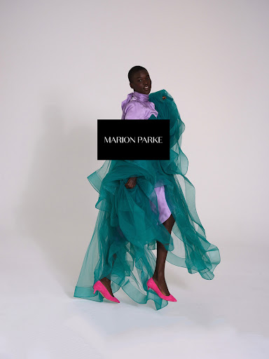 Fashionable Black woman poses in a lavender blouse, draped teal fabric, and pink heels against a white background in a studio shot.