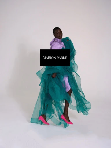 Fashionable Black woman poses in a lavender blouse, draped teal fabric, and pink heels against a white background in a studio shot.