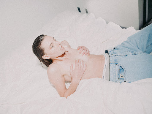 A topless woman lying on a white bed wearing light blue jeans with visible branded waistband, styled by Anna Katsanis.