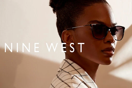 A woman poses wearing Nine West sunglasses and a plaid blazer in a studio, showcasing a fashion and beauty aesthetic.