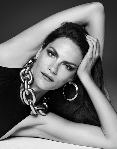 Black and white studio portrait of a woman with dark hair and heavy eye makeup, wearing large jewelry, lying down with a hand behind her head.