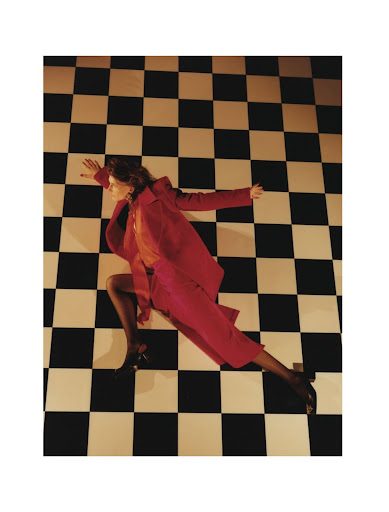 Fashion photograph featuring a woman in a red suit lying on a checkered floor, with her arms outstretched and legs bent.