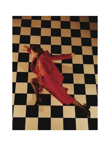Fashion photograph featuring a woman in a red suit lying on a checkered floor, with her arms outstretched and legs bent.