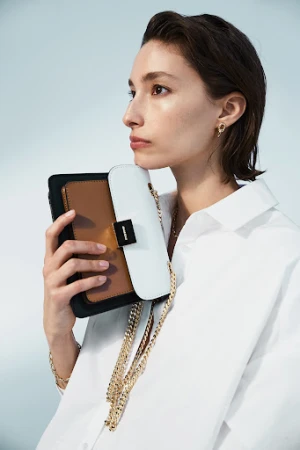 Woman in white shirt holding a structured color-block handbag with gold chain strap against a pale blue background