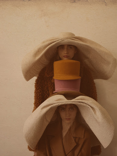 Three women are stacked vertically, each wearing a large hat. The top and bottom women wear cream-colored hats and the center wears two smaller hats.