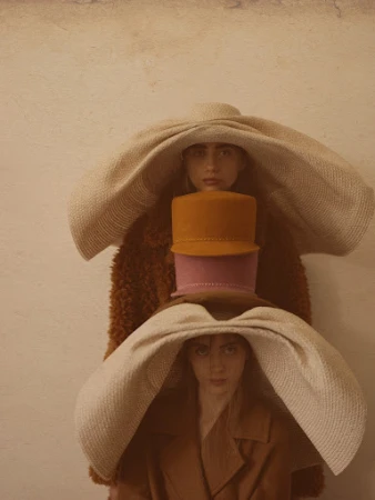 Three women are stacked vertically, each wearing a large hat. The top and bottom women wear cream-colored hats and the center wears two smaller hats.