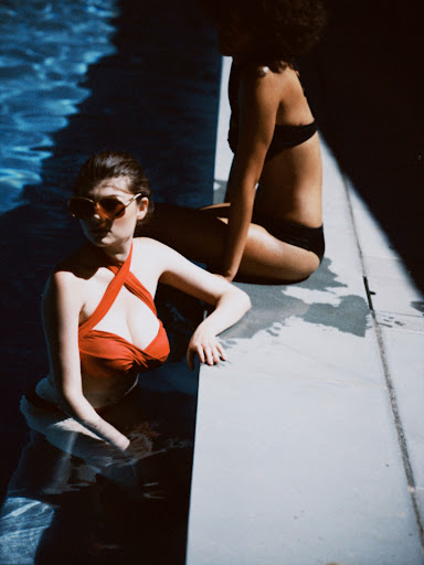 Two women in swimwear relax by a pool in bright sunlight. One woman is in the water and the other sitting on the edge.