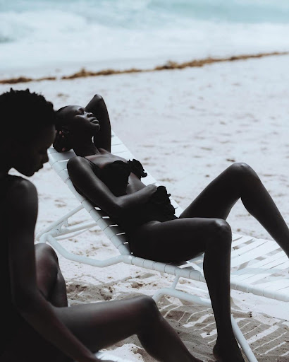 Two women in swimwear relax on a beach. One is reclining on a lounge chair while the other sits nearby, enjoying the ocean view.