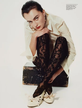 Woman wearing white blouse, black lace tights, and bow shoes crouching on a box against a white background