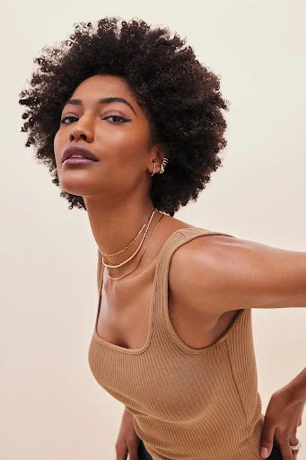 Close-up shot of an African American woman with a tan ribbed top, gold necklaces, and afro hair, looking confidently at the camera.