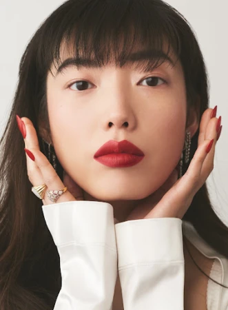 Close-up portrait of a woman with bold red lips and long red manicured nails framing her face against a seamless neutral background.