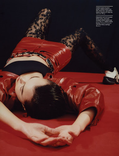 Woman in red leather jacket and black lace tights lying on a red surface against a dark background in a fashion editorial shot.