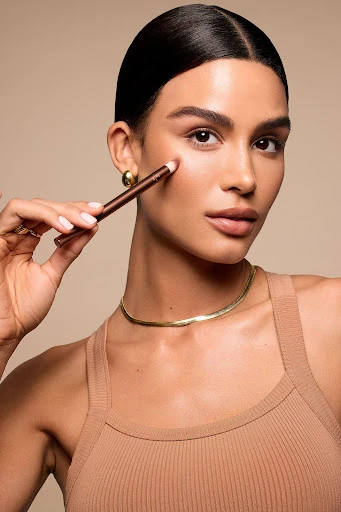 Close-up of a woman applying makeup with a brown eyeliner pencil. She has tan skin and a gold choker necklace.