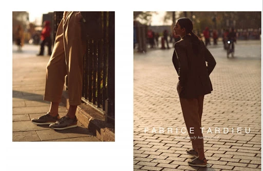 Fashion shot featuring a slender adult female model in tan pants and a dark blazer, posing in an urban setting with warm lighting.