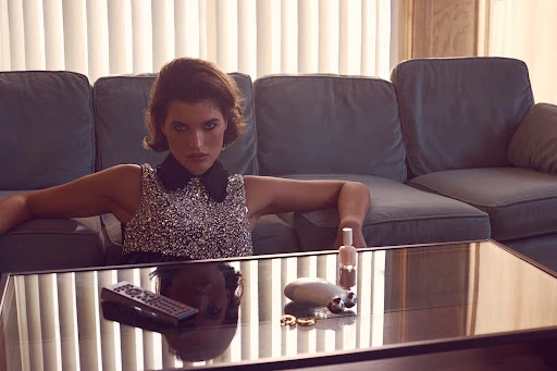 Woman in a sequin dress reclines on a blue couch in an indoor setting, with a coffee table and window blinds visible.