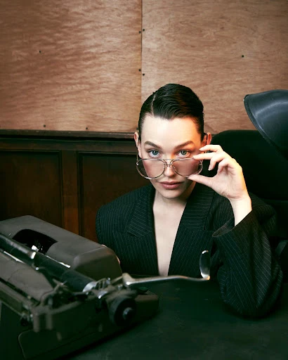 Woman in pinstripe blazer poses with antique typewriter, wearing clear eyeglasses in a wood paneled indoor environment.