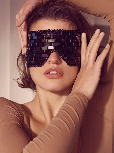 Woman wearing beaded sleep mask with hands near face in indoor setting. Beauty, fashion, and wellness concept with neutral colors.