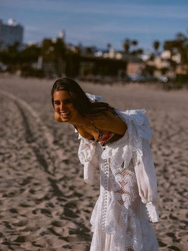 A woman on a beach smiles while wearing a white lace dress with a floral swimsuit top, with ocean and city buildings in the distance.