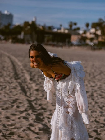 A woman on a beach smiles while wearing a white lace dress with a floral swimsuit top, with ocean and city buildings in the distance.