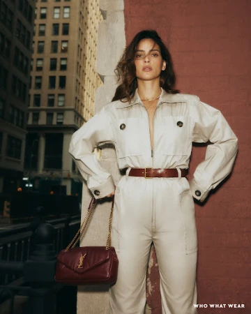 Woman in a beige utility jumpsuit and brown leather belt holding a burgundy quilted chain handbag outdoors