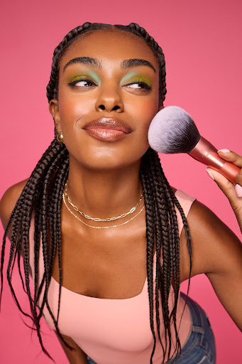 A Black woman applies makeup with a brush in a beauty setting, showcasing her complexion, hairstyle, and makeup artistry on a pink background.