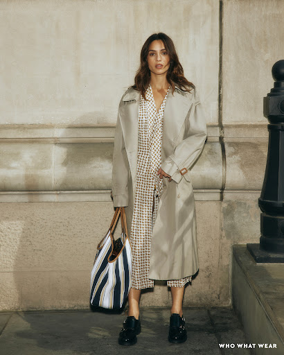 Woman wearing a beige trench coat over a patterned dress and holding a striped tote bag in an urban street setting.