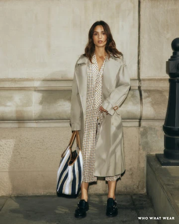 Woman wearing a beige trench coat over a patterned dress and holding a striped tote bag in an urban street setting.