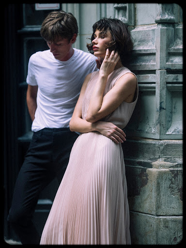A brunette woman in a pleated dress leans against a stone wall next to a man in a white t-shirt in an outdoor urban setting.