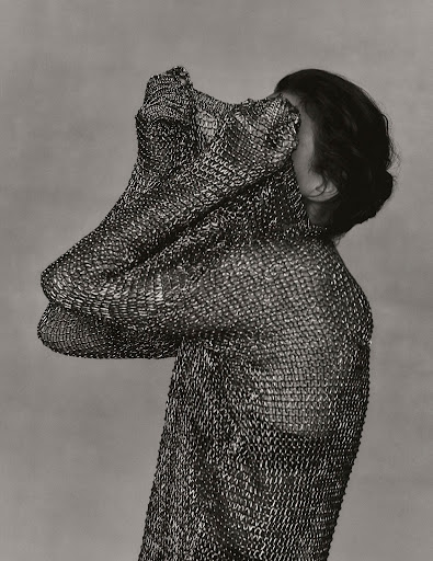 Monochrome portrait of a person draped in a textured metal mesh garment, partially covering the face, against a soft grey background.