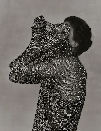 Monochrome portrait of a person draped in a textured metal mesh garment, partially covering the face, against a soft grey background.