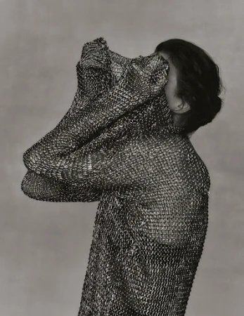 Monochrome portrait of a person draped in a textured metal mesh garment, partially covering the face, against a soft grey background.