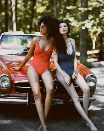 Two women in swimsuits pose near a classic red car in a lush, green outdoor setting. The image has a vintage feel.