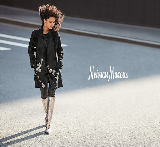 Woman with voluminous dark curly hair wearing an embroidered black coat and tall silver boots walking on an asphalt street