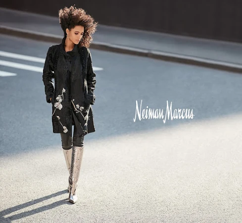 Woman with voluminous dark curly hair wearing an embroidered black coat and tall silver boots walking on an asphalt street