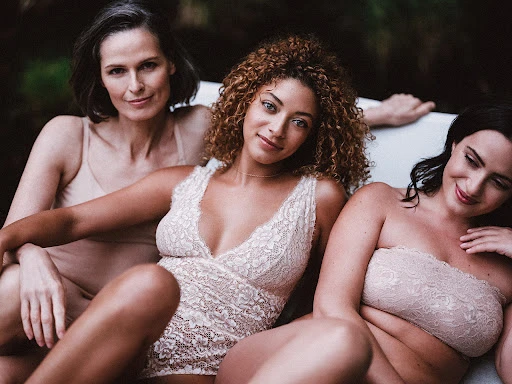 Three women wearing neutral beige lingerie posing together outdoors with greenery visible in the blurred background