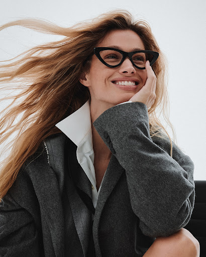 Smiling woman with long hair poses in glasses and an oversized grey wool coat against a white background in a portrait-style shot.