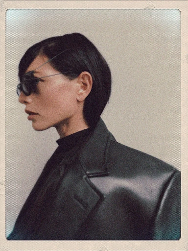 Portrait of a stylish adult female model in profile wearing dark sunglasses, a black turtleneck, and a black jacket against a neutral backdrop.