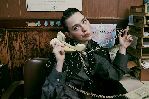 Stylish woman in a grey suit sits at a desk and poses with a vintage telephone in an indoor office environment with warm lighting.